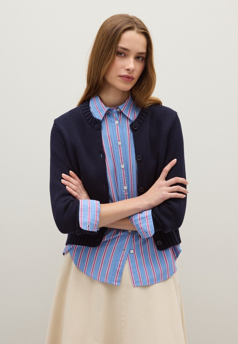 Cardigan lavorato a maglia blu navy sopra una camicia a bottoni a righe blu con righe rosa e bianche. Il cardigan ha un colletto rotondo e bottoni.