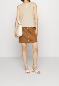 Beige ilman hihoja neulepaita V-päällä, ruskea tekosuedehame, jossa on etupainikkeet, valkoinen käsilaukku ja vaaleat mulesit paksuilla koroilla.