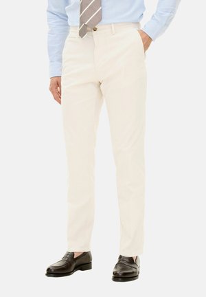 Pantalon beige clair en tissu lisse, avec une coupe slim, poches avant et taille mi-haute, associé à des mocassins foncés.