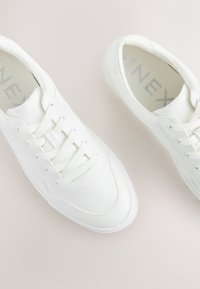 Baltos sportbačiai su lygiu sintetiniu viršumi, apvalia nosimi, su raišteliais ir textūruota guminė pado danga. Yra subtilūs siuvimo detalės.