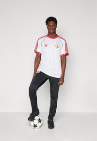 Witte voetbalshirt met rode accenten en geometrisch patroon, gecombineerd met zwarte broek en zwarte voetbalschoenen, staande op een voetbal.