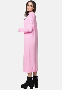 Langes, pinkes Strickkleid mit gerippter Textur und hohem Kragen. Kombiniert mit schwarzen, hohen Stiefeletten. Seitenansicht präsentiert.