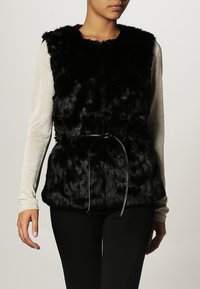 Musta tekoturkisliivi, jossa pehmeä pinta ja vyötäröllä solmittava nauha. Hihaton malli, käytetty kimaltavan pitkähihaisen paidan päällä.