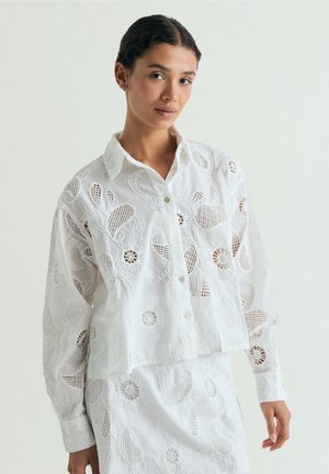 Mujer con camisa blanca bordada abotonada y falda a juego con patrones calados florales y de estilo paisley, de pie frente a un fondo liso.
