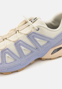 Light-toned sneaker featuring mesh and synthetic materials, purple accents, textured surfaces, and a distinct geometric design on the sides.