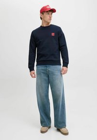 Mann, der zur Seite schaut, trägt eine rote Mütze, einen marineblauen Sweatshirt, weite hellblaue Jeans und beige Schuhe, steht vor einem schlichten hellen Hintergrund.