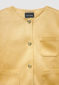 Gelbe Jacke aus Kunstwildleder mit rundem Halsausschnitt, goldfarbenen Knopfverschlüssen und einer Brusttasche. Glatte Textur, strukturierte Form, minimalistisches Design.