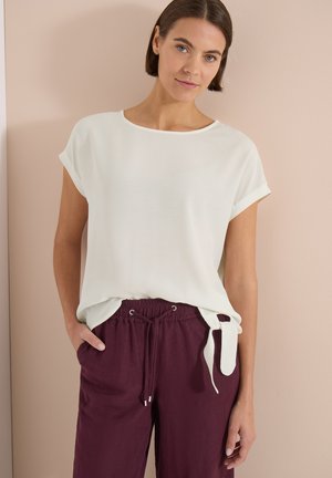 Frau trägt ein weißes kurzärmeliges Shirt mit seitlicher Schleife und bordeauxrote Jogginghosen, steht vor einer beigen Wand.