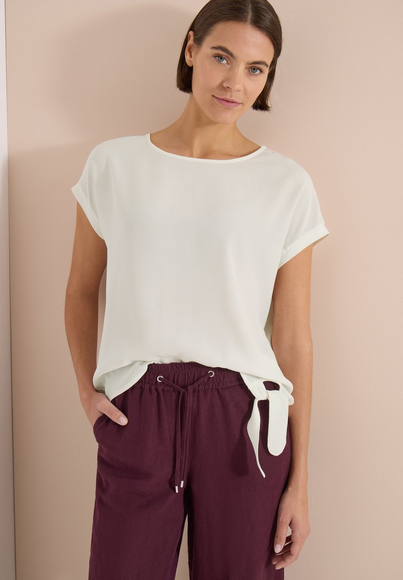 Femme portant un haut blanc à manches courtes avec un lien sur le côté et un pantalon bordeaux à cordon, debout contre un mur beige.