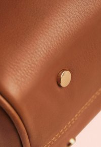 Sac en cuir marron avec une texture lisse, doté d'une fermeture à pression en métal doré et de coutures visibles le long du bord.