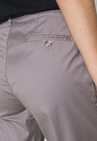 Grijze stoffen broek met een gladde textuur, voorzien van een achterzak met een zilveren knoop en een puntige flapdetail.