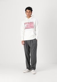 Hvit hettegenser med rosa logo-design, kombinert med mørkegrå løse bukser og hvite sneakers, stående på en ensfarget lys bakgrunn.