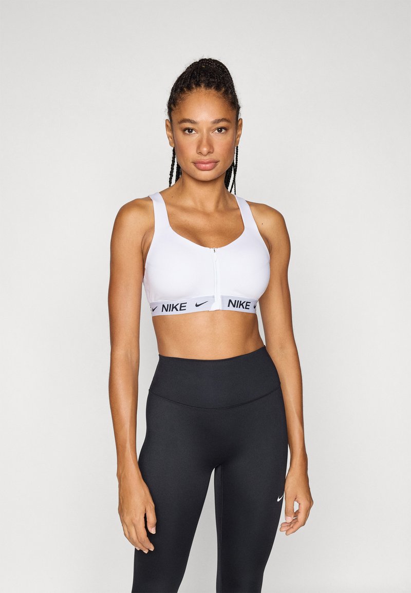 Witte sportbeha met ritssluiting aan de voorkant, dikke banden en een zwarte elastische band met merknaam. Gecombineerd met hoge zwarte leggings. Gladde stoftextuur.