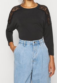 Maglietta nera a maniche lunghe con dettagli in pizzo sulle spalle, abbinata a jeans in denim blu chiaro a vita alta; tessuto morbido con vestibilità comoda.