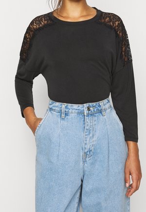 Schwarzes Langarmtop mit Spitzenakzenten an den Schultern, kombiniert mit hochgeschnittenen hellblauen Jeans; glattes Material mit lockerer Passform.