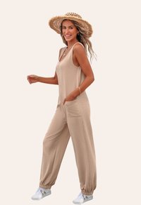 Beige mouwloze jumpsuit met zijzakken en elastische enkels, gemaakt van zachte stof, gecombineerd met witte sneakers en een strohoed.