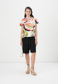 Cămașă scurtă, multicoloră, cu modele abstracte, asortată cu pantaloni scurți negri, croiți, și sandale cu brâu bej; ținând o geantă mică albă.