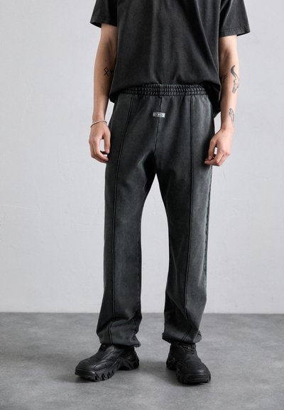 Pantaloni jogger neri con vestibilità rilassata, vita con coulisse, cuciture a contrasto e etichetta sul davanti. Indossati con scarpe da ginnastica nere.