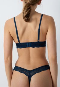 Soutien-gorge en dentelle bleu foncé et string assorti avec bretelles réglables, bords festonnés et motif floral ; fermeture à l'arrière visible sur le soutien-gorge.