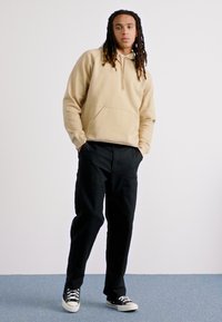 Beige capuchon sweatshirt met een voorzak, gecombineerd met zwarte losse broek en zwart-witte sneakers op een lichtblauwe vloer.