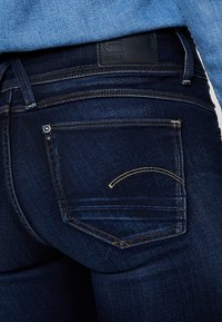 Jeans de mezclilla azul oscuro con un primer plano del bolsillo trasero que presenta un acento de costura amarilla y una etiqueta de cuero negra en la cinturilla.