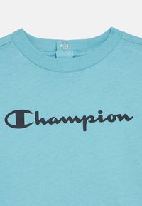 Ανοιχτό μπλε μπλουζάκι με στρογγυλή λαιμόκοψη και μαύρο λογότυπο "Champion" τυπωμένο στο στήθος. Απαλή κανονική βαμβακερή υφή. Κλείσιμο στο πίσω μέρος.