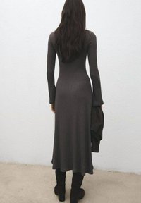 Vestido largo ajustado de color gris oscuro con detalle acanalado, mangas largas, bajo acampanado y material texturizado; combinado con botas oscuras hasta la rodilla.