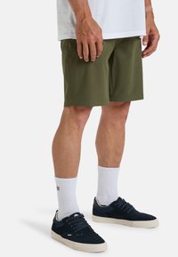 Pantaloni corti verde oliva, in tessuto leggero, taglio dritto, con tasche laterali. Indossati con sneakers blu navy scure e calzini bianchi a crew. Design semplice.