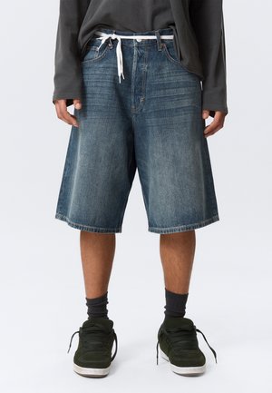 Person trägt lockere, kniehohe, verblasste Jeansshorts mit weißer Kordel, dunkelgraues Langarmshirt, schwarze Socken und dunkelgrüne Turnschuhe.