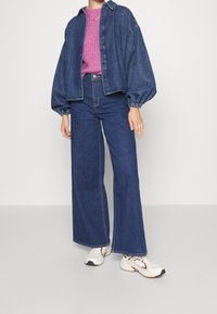 Veste en jean avec manches bouffantes portée sur un pull tricoté rose, associée à un jean en denim à jambes larges et des baskets blanches. Palette de couleurs bleu foncé.