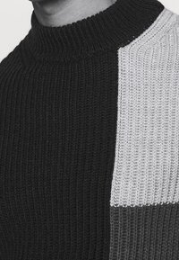 Stickad polotröja med ett svart och grått blockmönster, ribbad struktur och en åtsittande passform runt halsen.