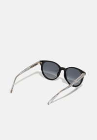 Lunettes de soleil noires avec monture ronde, verres dégradés foncés et branches transparentes fines avec des accents dorés.