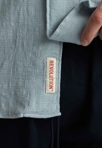 Lichtblauwe textuurstof met een geweven patroon, met een rode "REVOLUTION" label op een witte achtergrond. Donkere achtergrond zichtbaar.