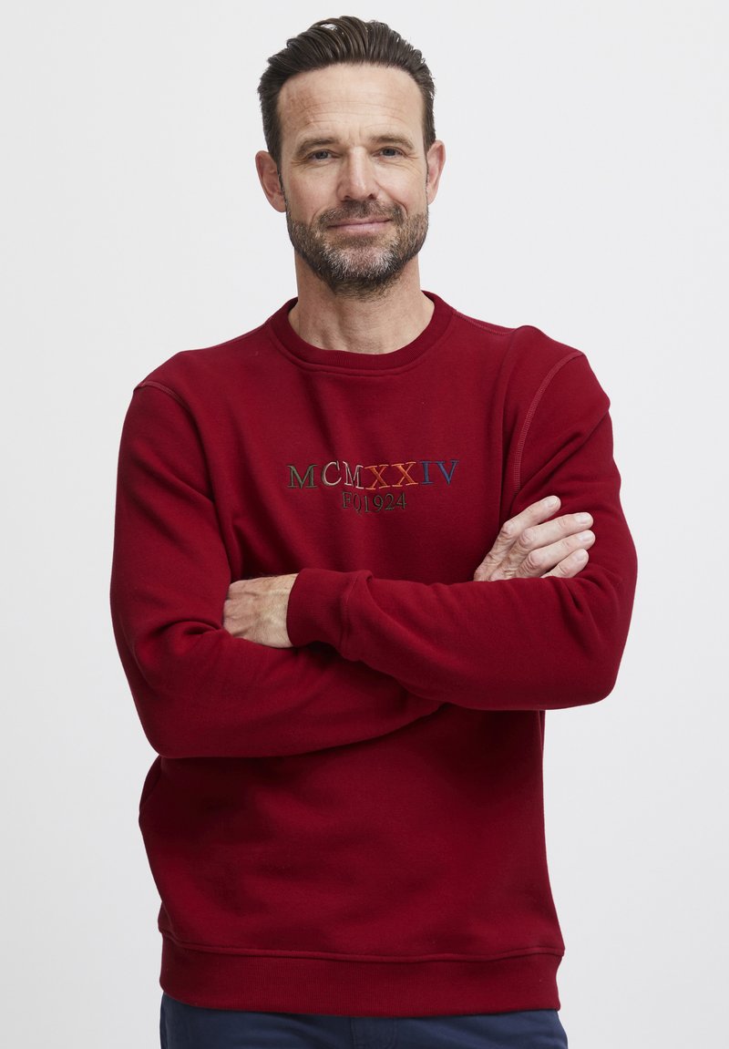 Červená mikina s kulatým výstřihem, s vyšitým textem v zelené, modré a oranžové. Vyrobena z měkké tkaniny s žebrovanými manžetami.