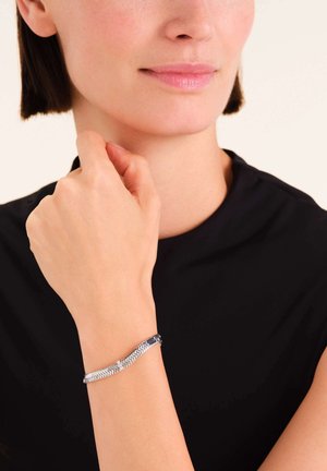 Pulsera de plata con un patrón en forma de chevrón, que presenta una pequeña piedra de acento, que se lleva en una muñeca de piel clara sobre una blusa negra.