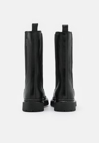 Mustat nahkaiset nilkkabotteet, joissa on sileä pinta, pystysuorat saumat ja paksut, tekstuuriset kumipohjat. Takana lenkkivetimet.