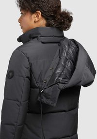 Woman with curly hair wearing a black padded jacket with an unzipped detachable hood, shown from the back and side.