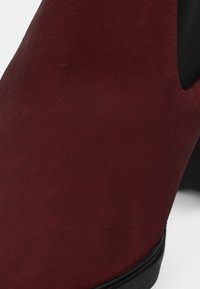 Stivale alla caviglia bordeaux con una morbida texture simile alla nubuck, caratterizzato da un design elegante, pannello laterale elastico e un tacco basso e arrotondato.