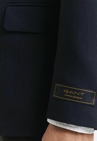 Mouw van een marineblauwe blazer met zichtbaar wit overhemdmanchet, met een zwart-gouden GANT American Sportswear label.
