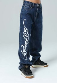 Jeans anchos de color azul oscuro con letras blancas de "Ed Hardy" a lo largo de la pierna izquierda. Zapatillas negras con acentos dorados y texto blanco. Textura clásica de mezclilla.