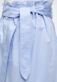 Jupe bleu clair taille haute avec une large ceinture en tissu nouée en nœud à la taille, montrant des coutures détaillées et des passants de ceinture.