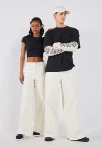 T-shirt noir court et pantalon large blanc portés par deux figures. Design de texte noir sur les manches longues et chaussures noires avec des semelles texturées.