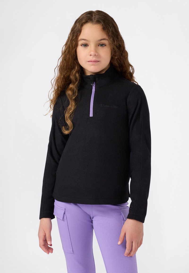 Ragazza con capelli lunghi e ricci, che indossa una felpa nera con zip e pantaloni cargo viola, in piedi davanti a uno sfondo bianco semplice.