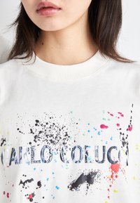 Hvid sweatshirt med ribbet halsudskæring, prydet med flerfarvede sprøjtede mønstre og en fremtrædende sort grafisk tekst "CARLO COLUCCIO."