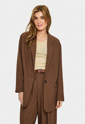 Vrouw met lang lichtbruin haar, gekleed in een losse bruine blazer en bijpassende broek over een beige top, staand tegen een effen witte achtergrond.