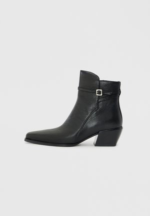 Bottines en cuir noir avec un bout pointu, un petit talon carré et une sangle décorative avec une boucle en argent. Texture lisse.