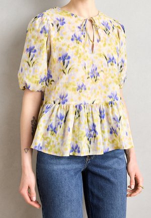 Blusa floral con mangas cortas abullonadas, dobladillo tipo peplum, pequeño escote en forma de ojo de cerradura con lazo frontal, combinada con vaqueros azules en una persona con tatuajes en los brazos.