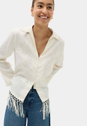 Smilende person iført en hvid skjorte med knapper, blomsterbroderi og frynser, stående med hænderne bag ryggen iført blå jeans.