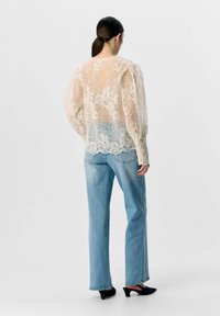 Blondebluse i beige med blomster mønstre, gennemsigtig stof, ballonærmer og en bølget kant, parret med lyseblå denim jeans og sorte hæle.