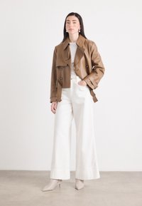 Een jonge vrouw staat zelfverzekerd in een bruine cropped jack en witte wijde broek, accessorized met statement oorbellen en beige hakken.